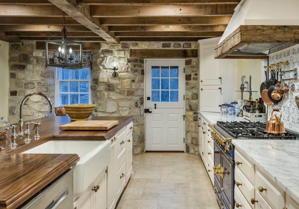 A cozy kitchen featuring rustic charm with exposed wooden beams and a combination of stone and white cabinetry. A large, white farmhouse sink is integrated into a wooden countertop, which holds a brown ceramic bowl and a cutting board. On the right side, a navy blue stove contrasts with the white marble countertop, adorned with various copper pots and utensils. A large window allows natural light to filter in, complemented by a simple black chandelier hanging above. A white door with glass panels provides access to the outside.
