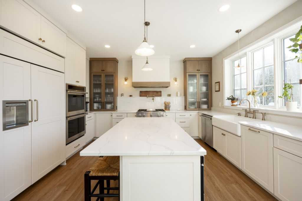 Bright, modern kitchen with white cabinets, marble island, wooden floors, farmhouse sink, stainless steel appliances, pendant lights, and large windows letting in natural light.