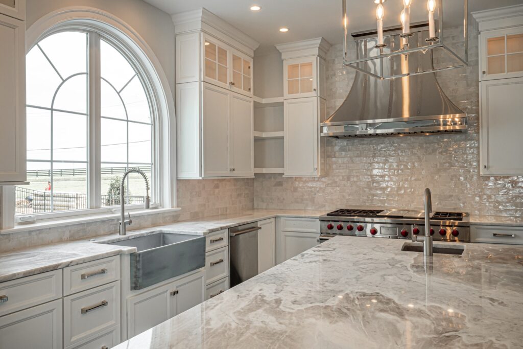 A modern kitchen featuring sleek white cabinetry with glass front details, a large marble countertop with a subtle gray pattern, and a farmhouse-style sink. The kitchen includes a stainless steel range with red knobs and a polished metallic exhaust hood above. A spacious arched window allows natural light in, revealing a view of greenery outside. Pendant lighting hangs above the island, complementing the contemporary design.