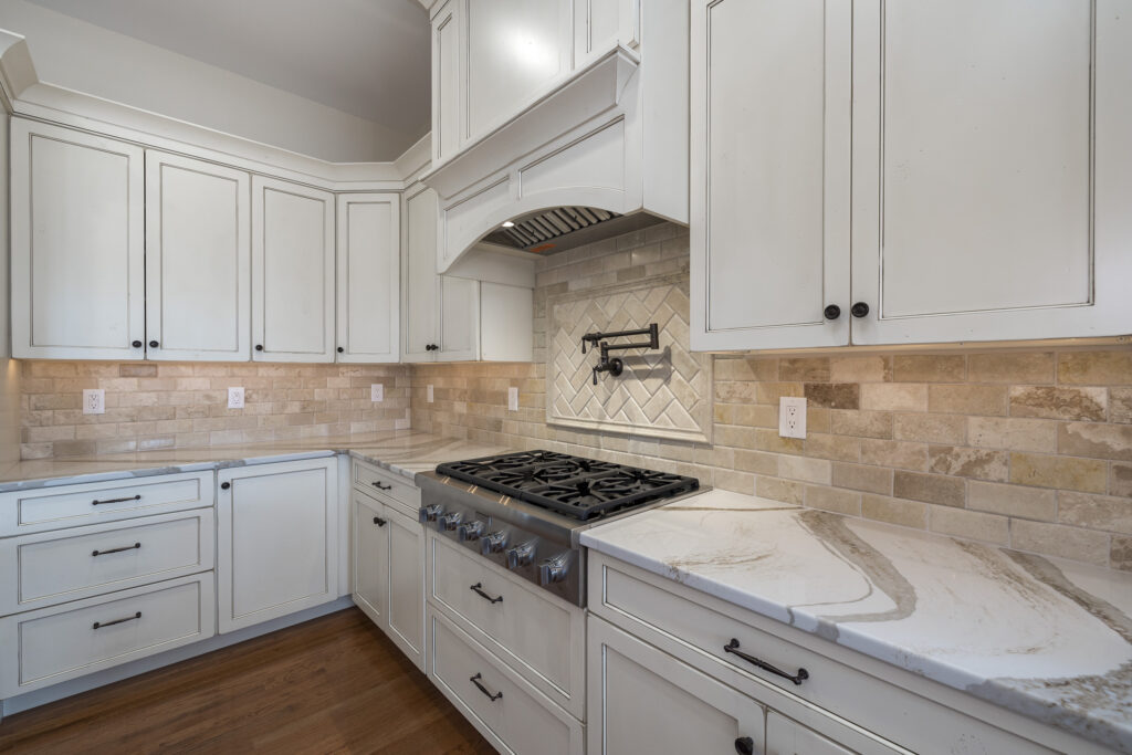 A modern kitchen featuring white cabinetry with black handles. The cabinets are complemented by a light-colored stone backsplash with a herringbone pattern and several electrical outlets. A spacious countertop with subtle veining extends along the base, supporting a large gas stove with multiple burners. The overall design conveys a clean and contemporary aesthetic.