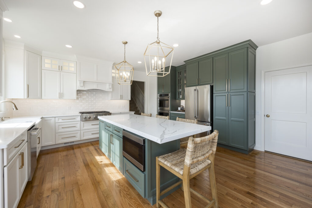 A modern kitchen featuring a large island with a white marble countertop and teal cabinetry. The island includes an built-in oven and a seating area with woven bar stools. White cabinets line the walls, complemented by a tile backsplash. Overhead, two gold geometric pendant lights illuminate the space. A stainless steel refrigerator and built-in ovens are visible on one side, while warm wooden flooring adds a cozy touch. Natural light fills the room, enhancing the clean and welcoming atmosphere.