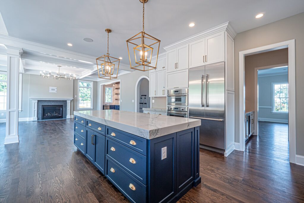 A modern kitchen featuring a large central island with a white marble countertop and dark blue cabinetry. Above the island, two geometric gold pendant lights hang. The kitchen has white cabinets and stainless steel appliances along one wall. A doorway on the right leads to another room, while large windows illuminate the space, showcasing a wooden floor and a cozy fireplace in the background.