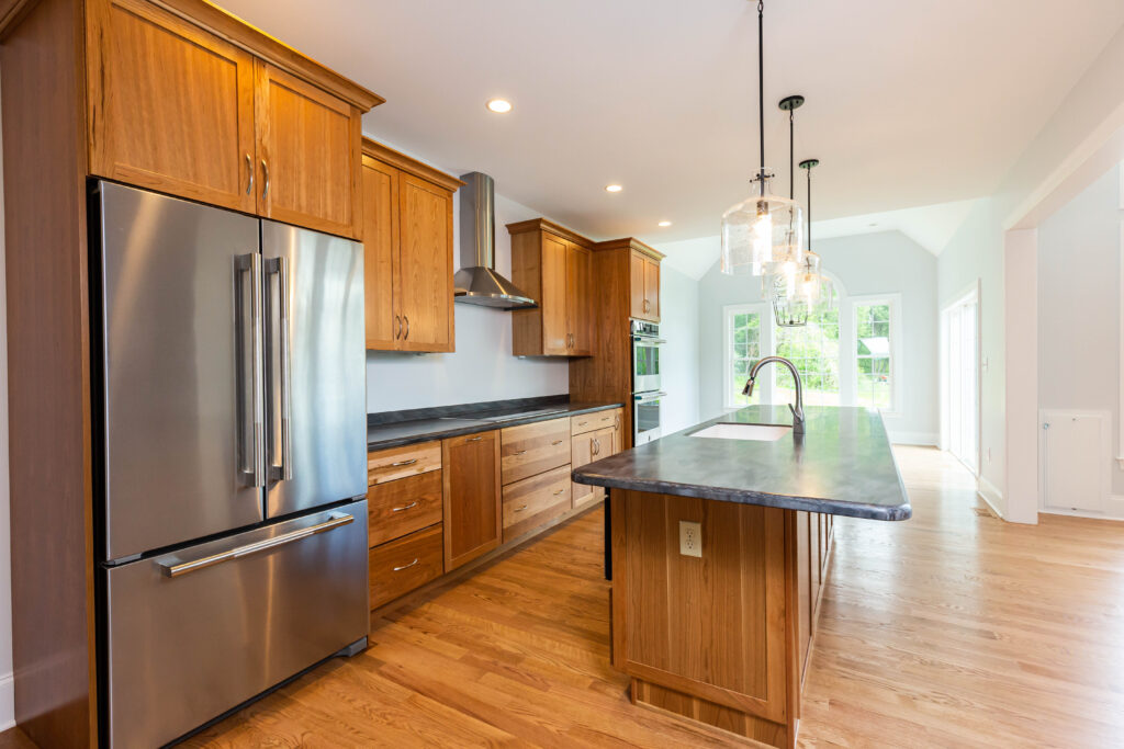 A modern kitchen featuring wooden cabinetry with a warm finish and a stainless steel refrigerator. The kitchen island has a dark countertop and a sink, while pendant lights hang above. A range hood is installed above the stove, and there are built-in ovens on one side. Natural light floods the space through large windows, highlighting the hardwood floor. The walls are painted a light color, contributing to an airy and inviting atmosphere.