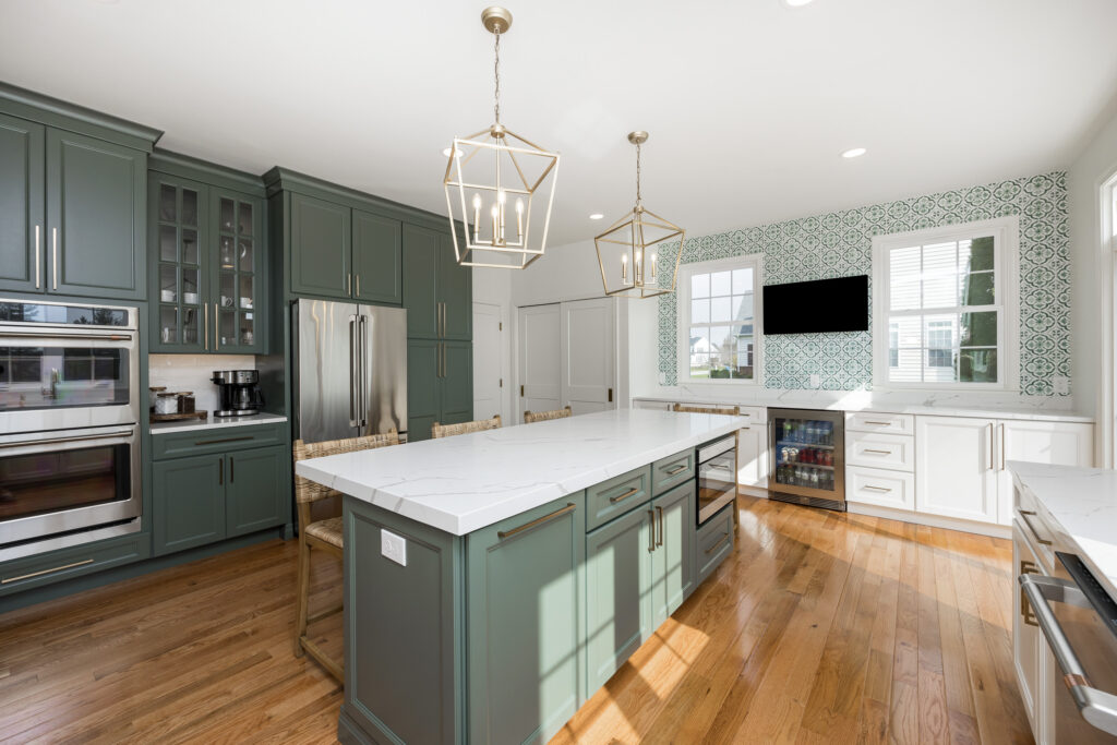 A modern kitchen featuring green cabinetry and a white marble countertop island. Two geometric pendant lights hang above the island, illuminating the space. The kitchen includes stainless steel appliances, including a refrigerator and an oven, alongside a coffee maker. A patterned backsplash complements the cabinetry. Natural light floods in through several windows with white blinds, and there is a small sitting area with wooden chairs. A wall-mounted television is visible in the room. The flooring is made of polished wood, adding warmth to the contemporary design.