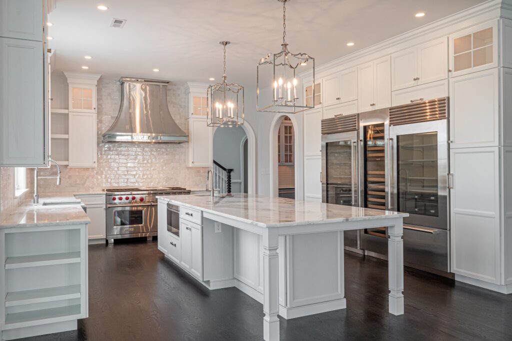 A modern kitchen featuring white cabinetry with glass-paneled doors, a large marble-topped island, and stainless steel appliances, including a double oven and a sleek hood above a gas range. The space is well-lit with three pendant lights hanging over the island and recessed lighting in the ceiling, creating a bright and welcoming atmosphere. The dark hardwood floor contrasts elegantly with the light cabinetry and countertops. A doorway leads to another room in the background.