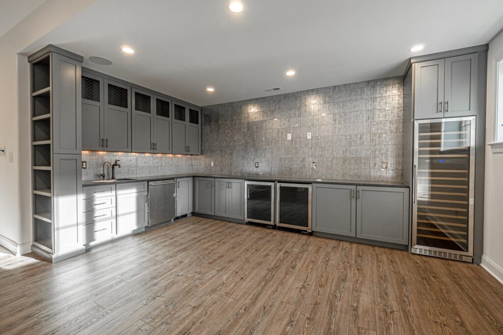 A modern kitchen features gray cabinetry with glass panels, complemented by a textured gray tile backsplash. The countertop is dark, with various built-in appliances, including a wine fridge and a stainless steel refrigerator. The space is well-lit by recessed lights, and the floor is made of warm-toned wood planks. The overall design is sleek and stylish, providing a contemporary aesthetic.
