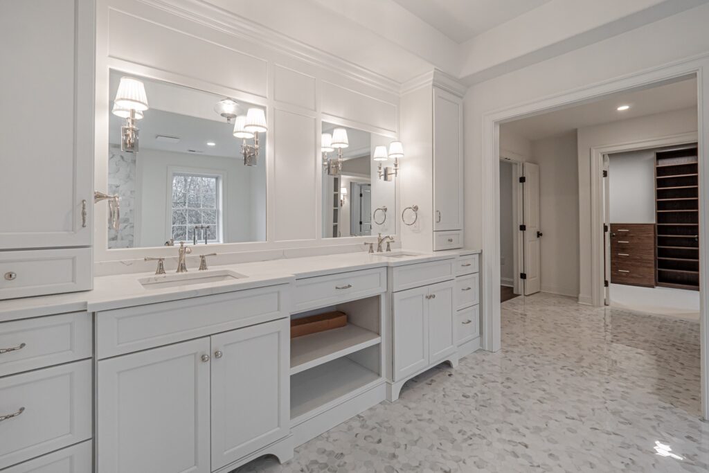 A bright and spacious bathroom featuring a double vanity with a white marble countertop and stylish silver faucets. Above the vanity are three large mirrors flanked by elegant wall sconces that provide warm lighting. The cabinetry is white, with multiple drawers and a partially open shelf displaying a wooden box inside. To the right, an open door leads to a closet with wooden shelving, and a large window allows natural light to fill the room. The floor is composed of light-colored, flat stones, enhancing the modern aesthetic.