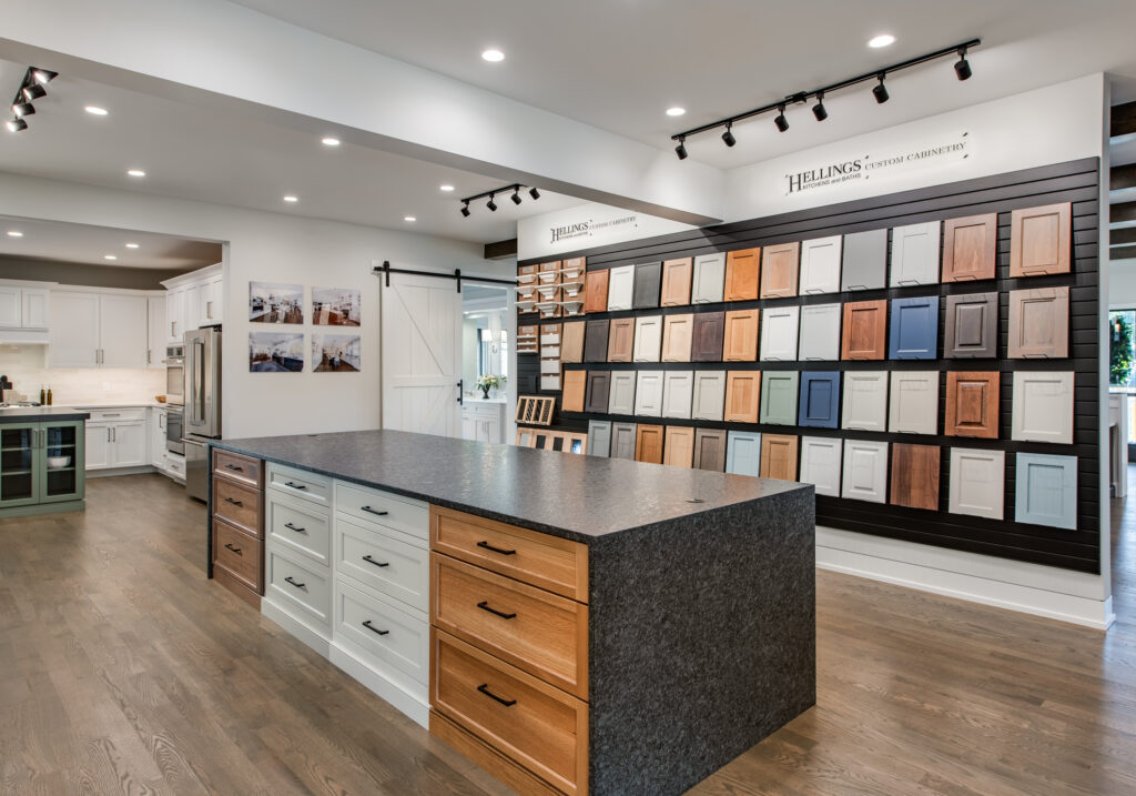 A modern open-concept kitchen and living area featuring a large island with a wooden countertop and a stainless steel sink. The kitchen has light gray cabinetry and a five-burner gas stove. Above the island, three round pendant lights hang. To the right, a cozy living area includes a gray sectional sofa, a light armchair, and a coffee table. Large windows allow natural light to fill the space, highlighting the wooden flooring. A staircase is visible in the background, leading to an upper floor.
