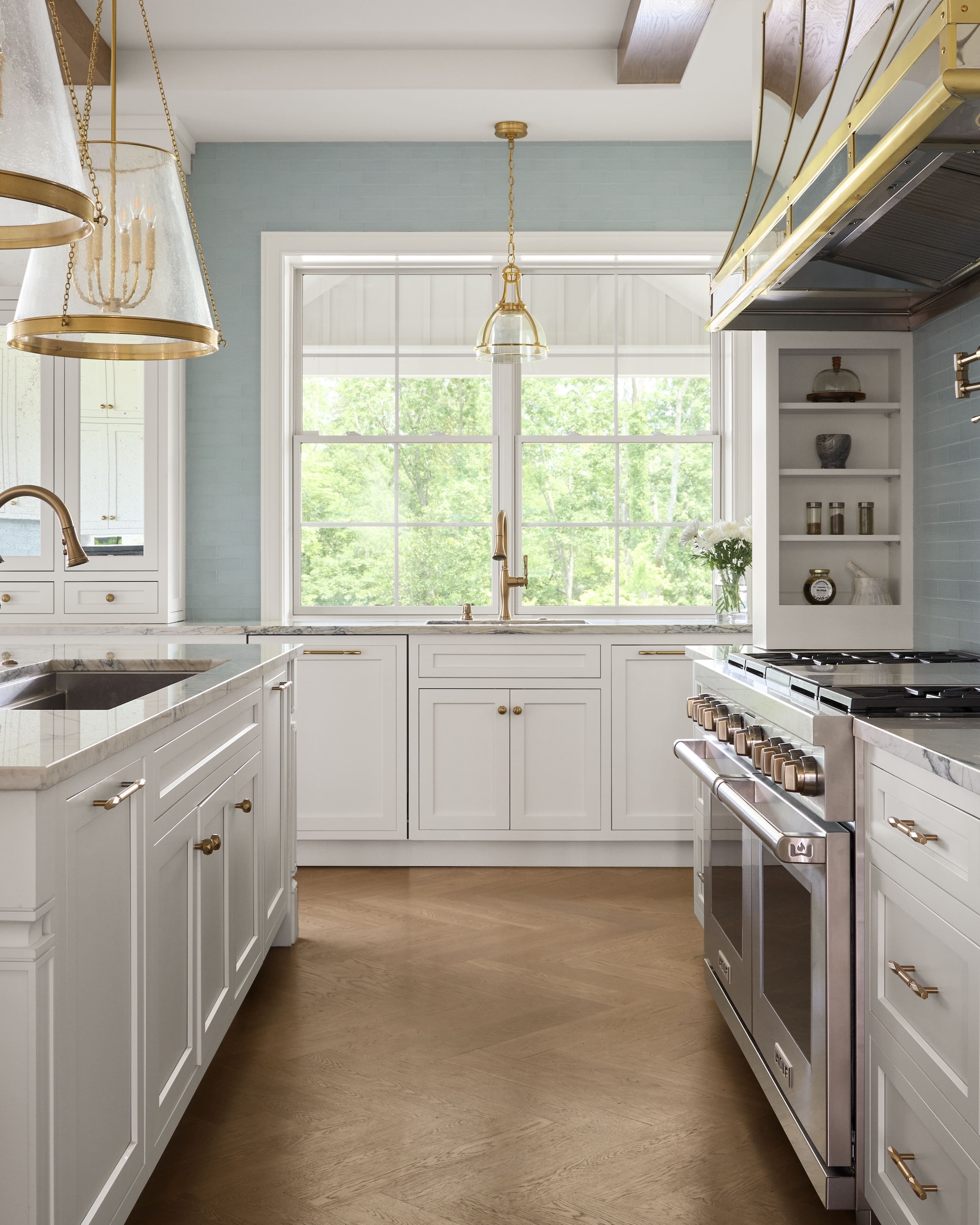 Bright, modern kitchen with white cabinets, gold hardware, marble countertops, a large stove, and two pendant lights. A large window above the sink lets in natural light, and the floor is a light wood.