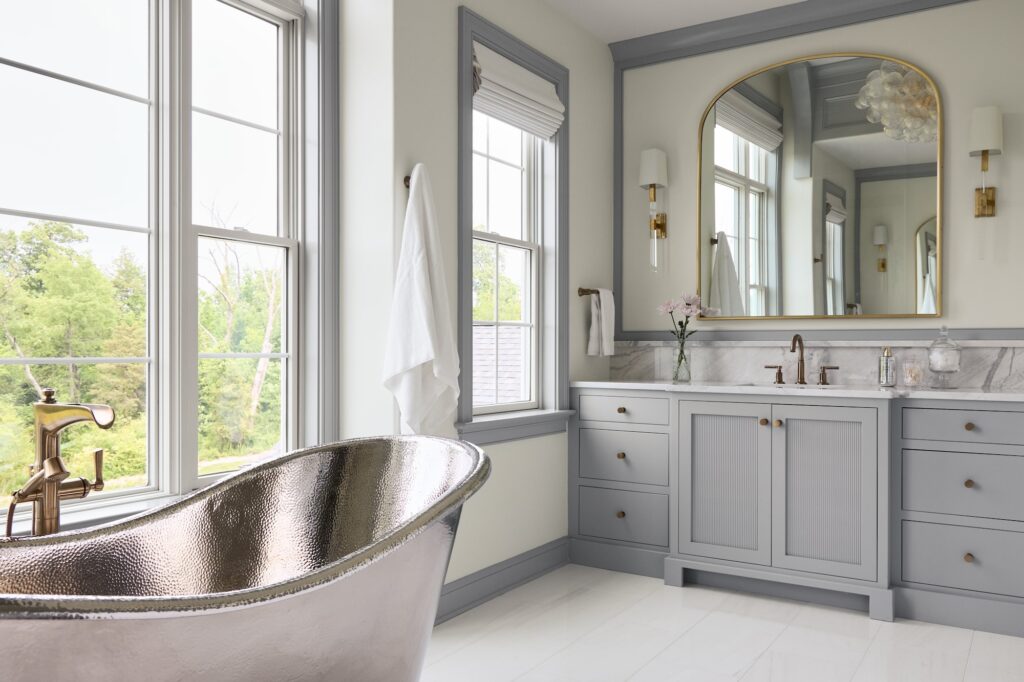 A bright, elegant bathroom with a metallic freestanding bathtub, gray cabinets with marble countertops, a large arched mirror, sconces, and tall windows letting in natural light and views of greenery outside.