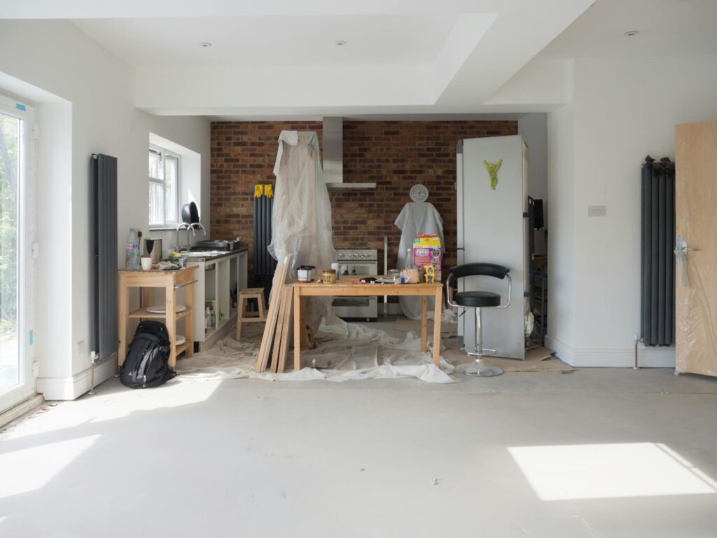A kitchen under renovation with exposed brick walls, covered appliances, scattered tools, paint tubs, and a floor partly protected by sheets. Some cabinets and a fridge are covered with cloths or plastic.