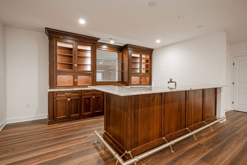Modern home bar with dark wood cabinets, glass shelves, a marble countertop, and a foot rail. The space has wood flooring, recessed lighting, and plain white walls, creating a clean and elegant look.